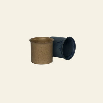 The ves Kura Cup Set of 2 by ves features one tan upright and one blue cylindrical cup, adding handcrafted ceramic charm to your decor, shown against a beige background.