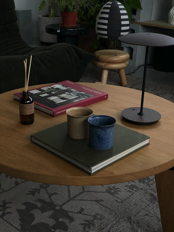 A round wooden coffee table from the ves Kura Collection displays a ves Kura Cup set of 2, a closed book, reed diffuser, and black lamp. In the background: a black chair, small stool, and potted plants complete the decor.